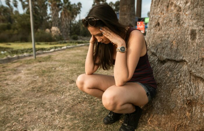 Ağaca yaslanmış, dizlerinin üstünde çökmüş, başını ellerinin arasına almış yorgun kadın; ruhsal yorgunluk, tükenmişlik sendromu, içsel çöküş, spiritüel yorgunluk